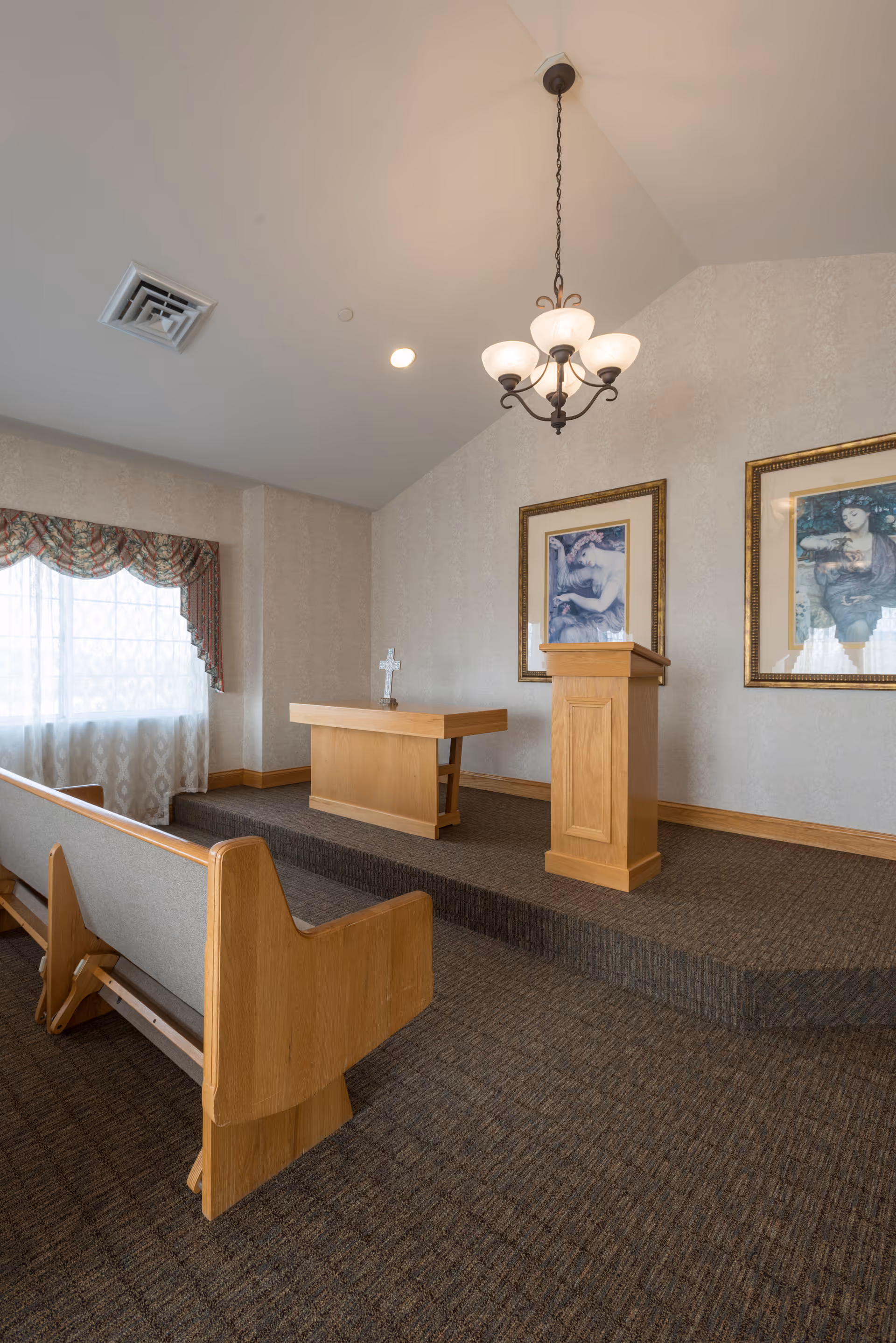 Interior of a small chapel or prayer room with wooden pews, a raised platform with a wooden altar and lectern, a cross on the altar, two framed paintings on the wall, a window with sheer curtains and a valance, and a hanging chandelier light fixture.