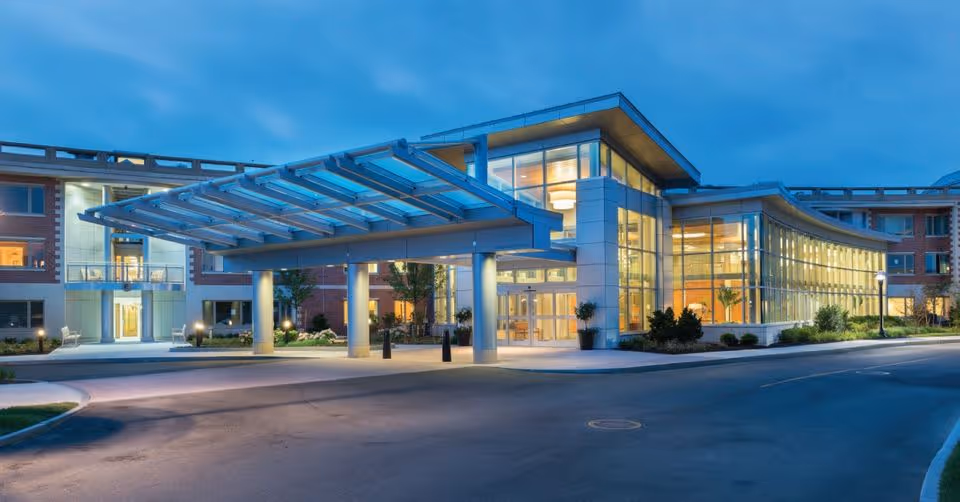 Exterior view of North Hill Retirement Community building at dusk, featuring a modern entrance with large glass windows and a covered drop-off area illuminated by lights.