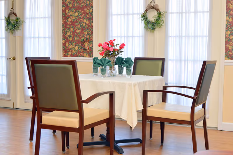 A dining area with a square table covered with a white tablecloth, set with four green folded napkins in glasses and a vase of red flowers in the center. Four wooden chairs with green and beige cushions surround the table. The room has large windows with sheer white curtains and floral wall decorations with hanging greenery.