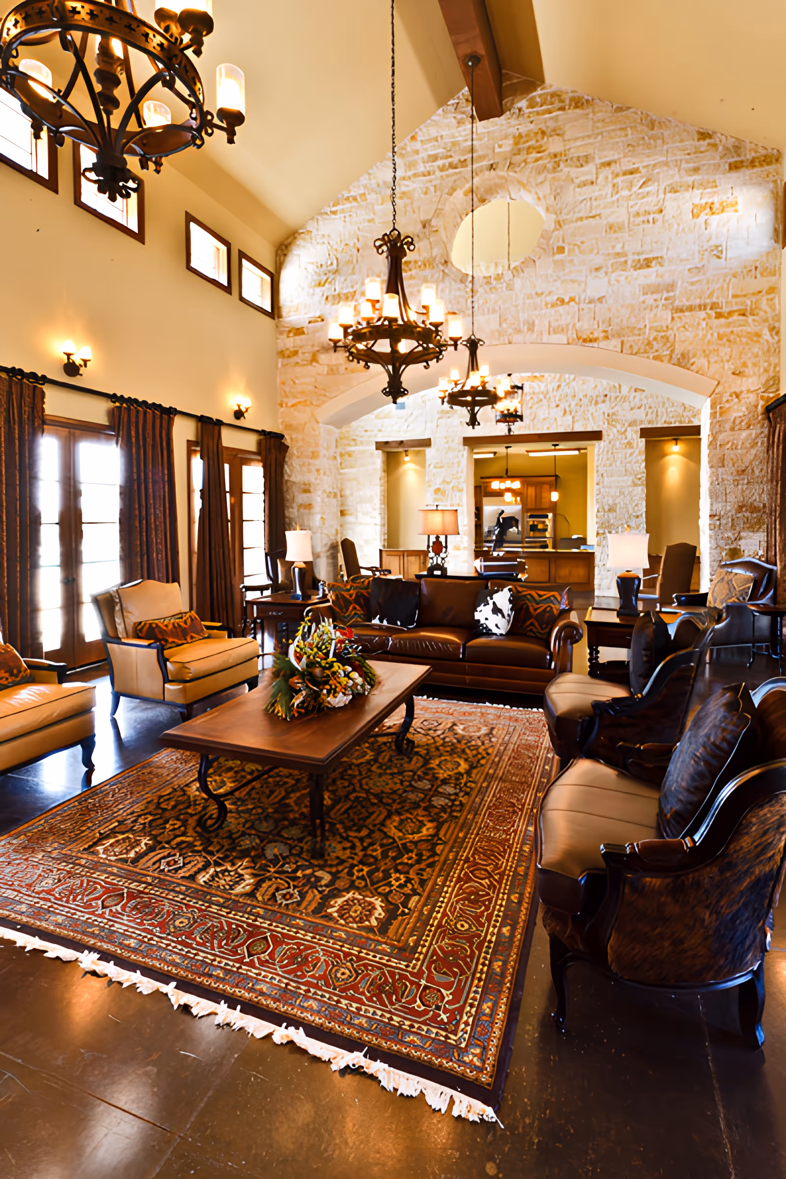 A spacious and elegant living room with high vaulted ceilings and stone walls. The room features a large patterned area rug, a wooden coffee table with a floral arrangement, leather sofas, and armchairs. Multiple chandeliers hang from the ceiling, and large windows with curtains allow natural light to fill the space. In the background, there is a view into a kitchen area.