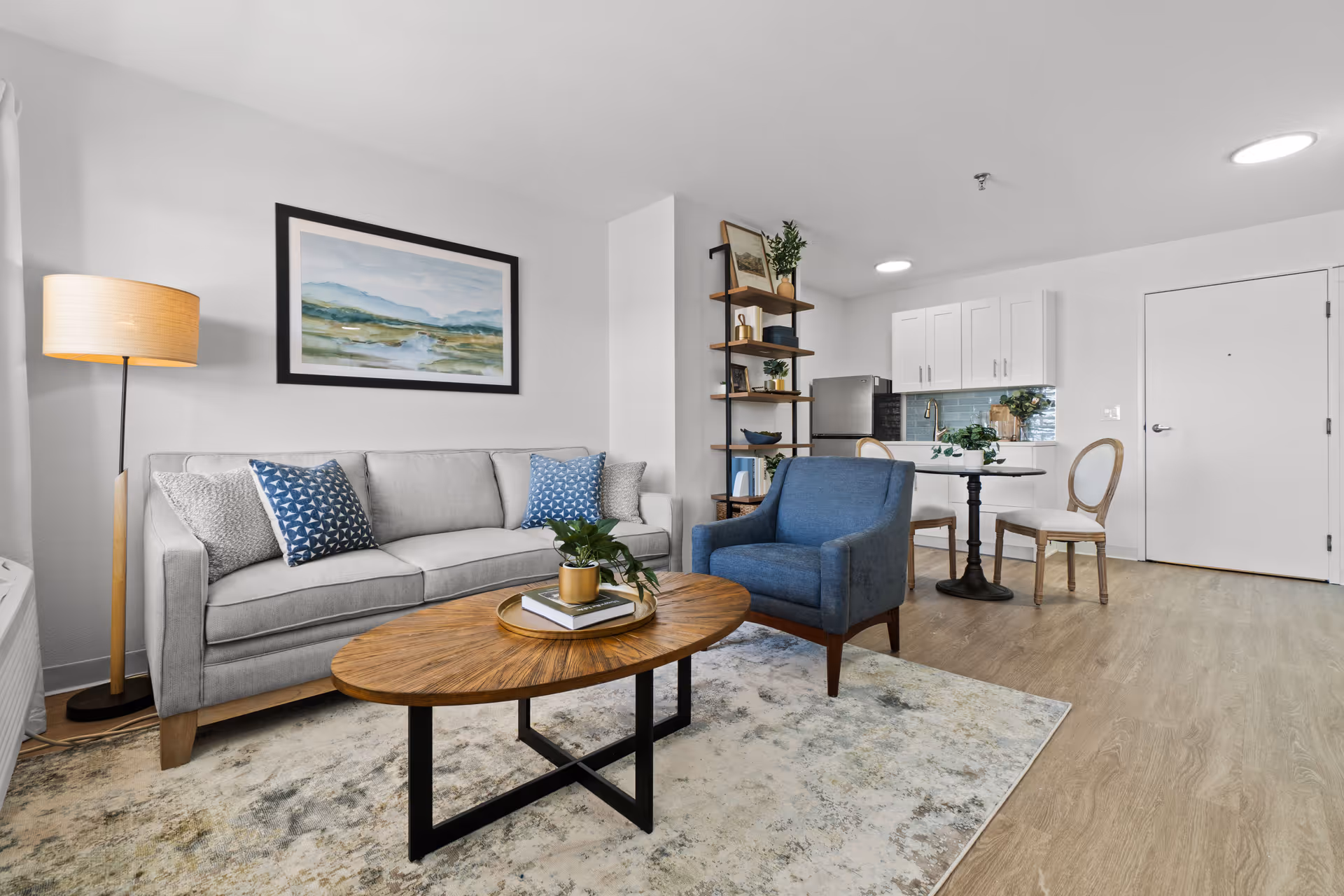 Bright modern living room with a gray sofa, blue armchair, wooden coffee table, and a small dining area by a kitchenette.