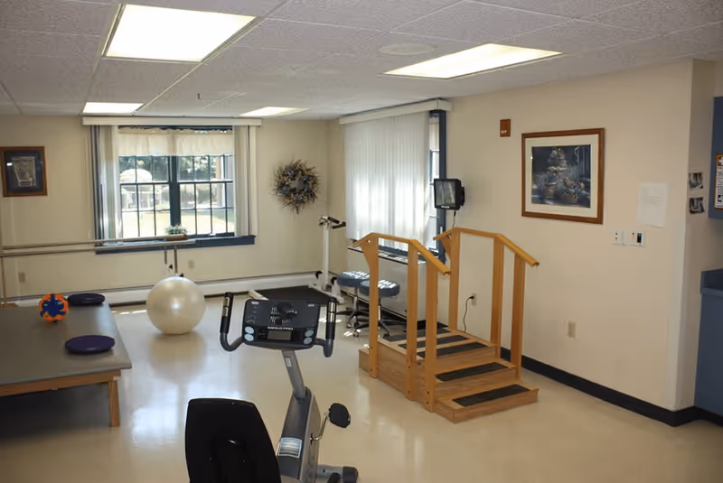 A small physical therapy or exercise room with a stationary bike, wooden stairs with handrails, a large exercise ball, balance cushions, and parallel bars near a window with vertical blinds. The room has beige walls, a framed picture, and fluorescent ceiling lights.