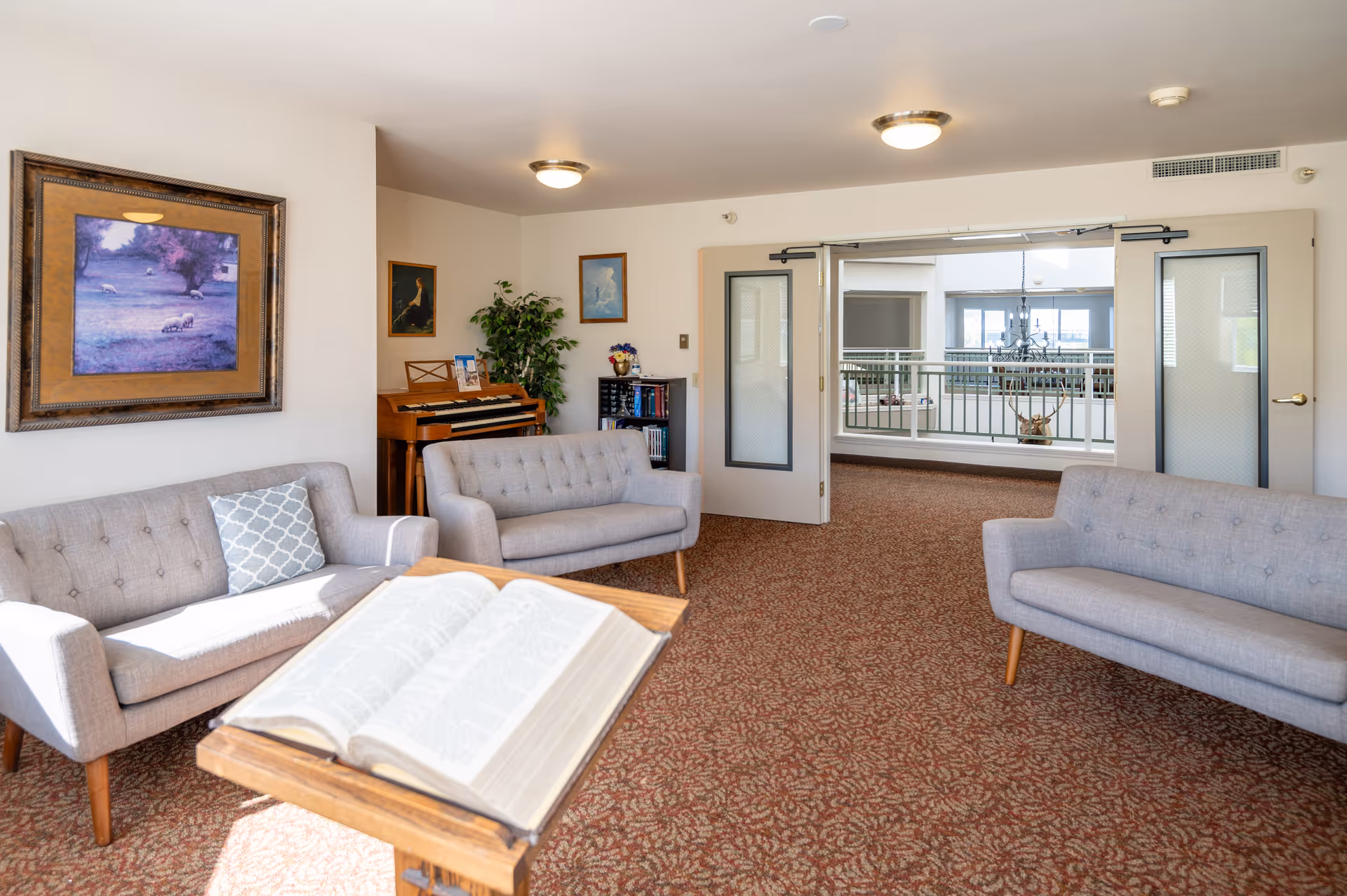 A bright senior living lounge with gray sofas, a piano, an open book on a stand, and double doors opening to a balcony area.