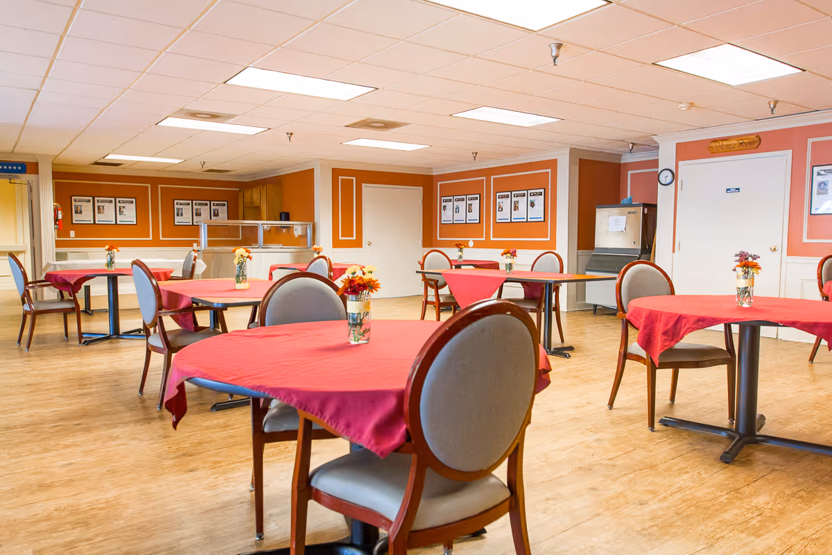 A dining room in a nursing center with several round tables covered with red tablecloths, each table having a small vase with flowers. The room has wooden flooring, orange walls with white trim, and framed pictures on the walls. There is a serving counter and an ice machine in the background.