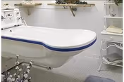 A white bathtub with a blue trim in a bathroom setting. The bathtub is mounted on metal legs and has a control panel on one end. The floor is covered with small mosaic tiles, and there are shelves on the wall holding towels and other bathroom items.
