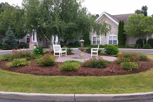A landscaped circular garden area with a central stone fountain surrounded by two white wooden chairs, various bushes, and trees. In the background, there is a single-story building with stone and siding exterior walls, windows, and an American flag near the entrance.