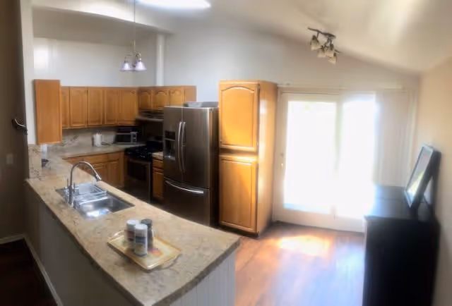 Bright kitchen with wooden cabinets, granite countertops, a double sink, stainless steel refrigerator, stove, microwave, and a small dining area near glass doors letting in natural light.