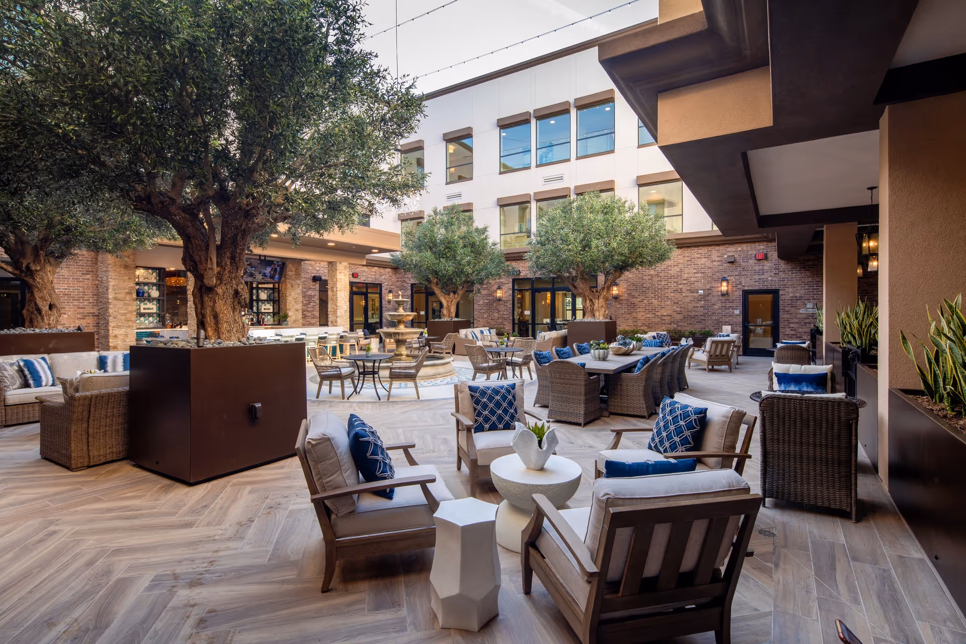 A spacious outdoor courtyard area with comfortable seating including cushioned chairs and sofas arranged around small tables. There are large potted trees and a central multi-tiered water fountain. The courtyard is surrounded by a multi-story building with large windows and brick walls, creating a relaxing and inviting atmosphere.