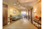 Well-lit nursing home common area with sofas, chairs, side tables, artwork, and overhead track lighting.