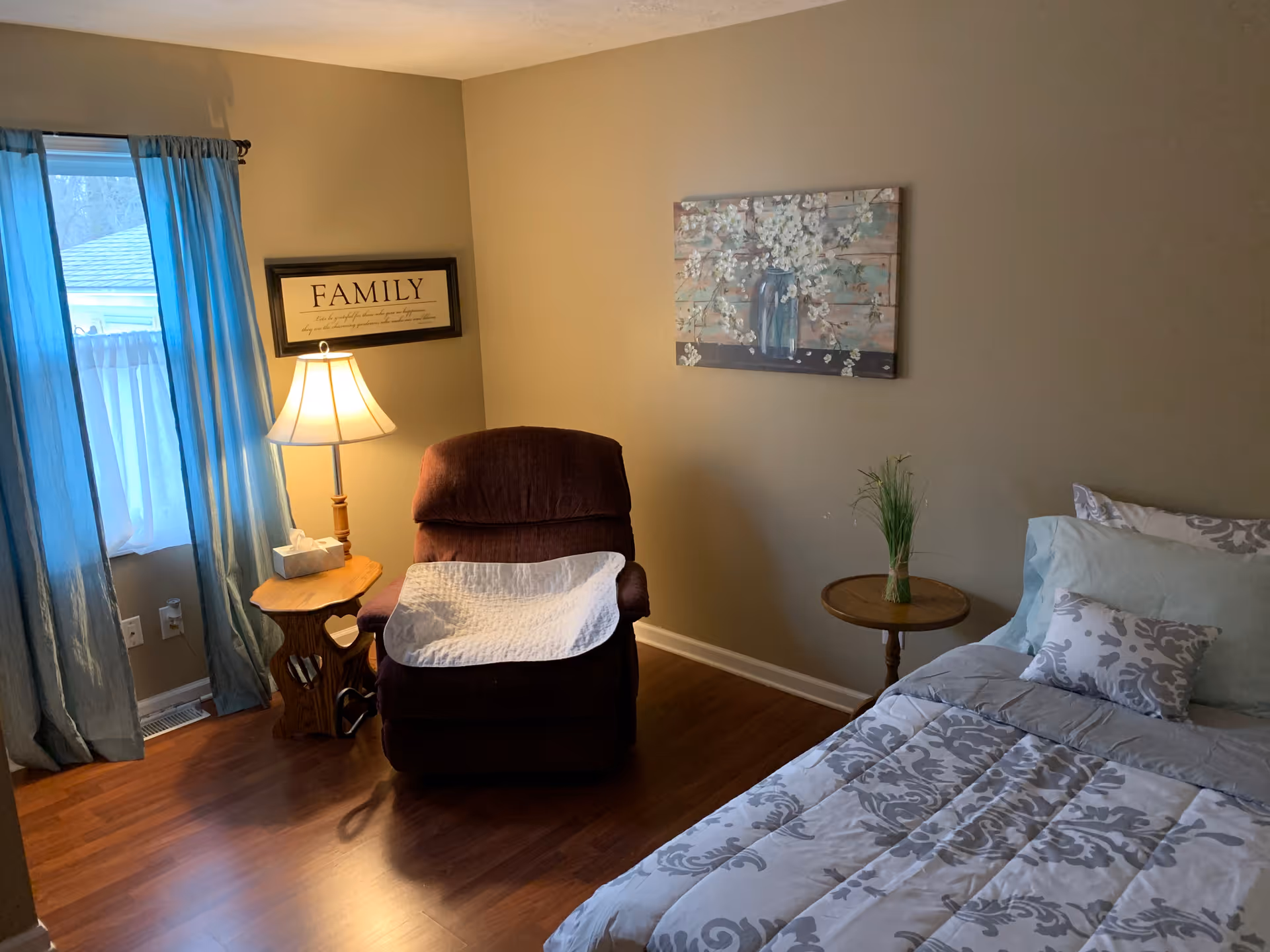 Cozy bedroom with a bed, recliner chair, lamp and side tables beneath wall art and a curtained window.