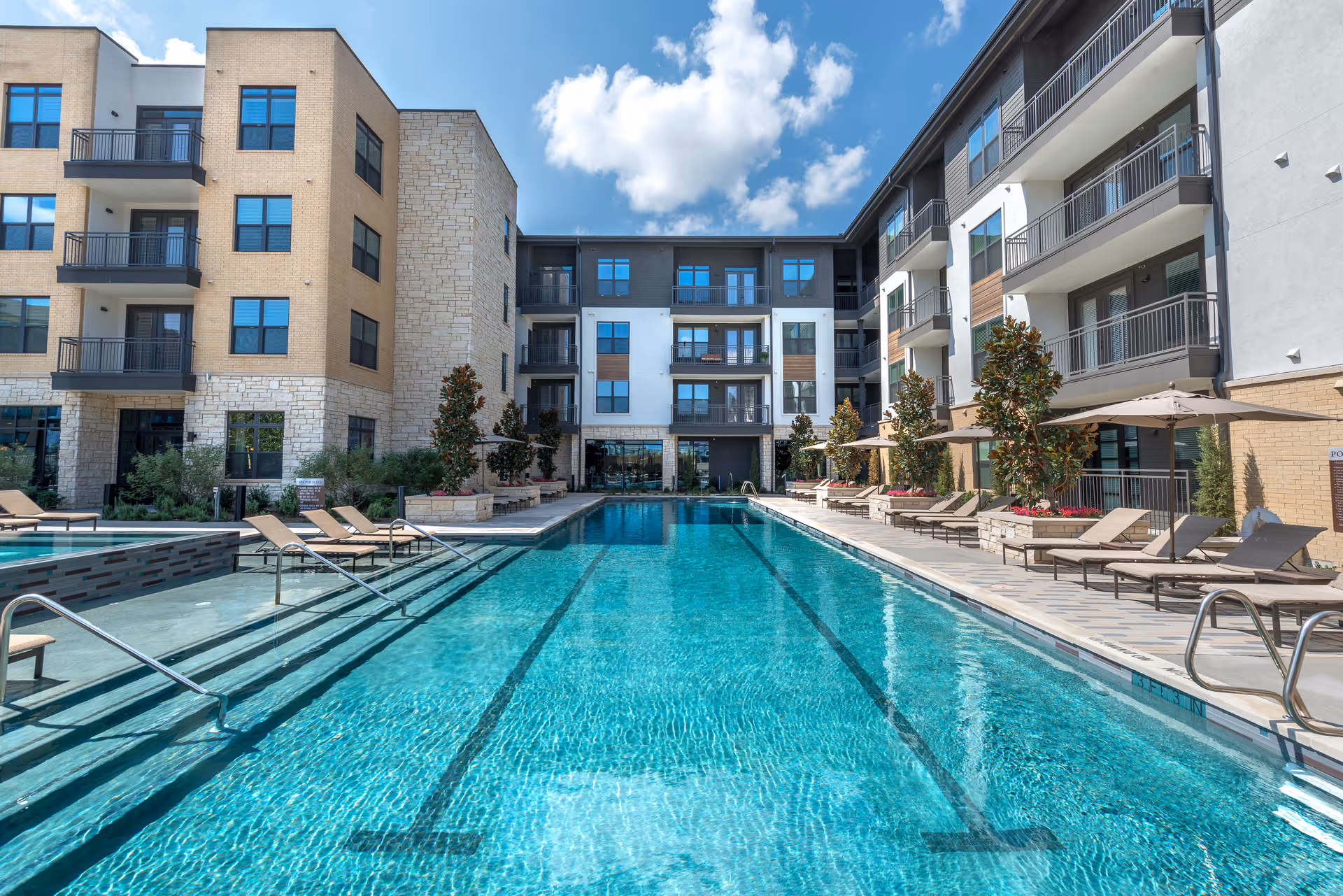 Outdoor swimming pool with clear blue water surrounded by lounge chairs and umbrellas, flanked by multi-story residential buildings under a partly cloudy sky.