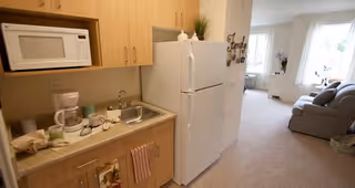 A small kitchen area with wooden cabinets, a microwave, coffee maker, sink, and a white refrigerator. The kitchen opens into a living room with a gray sofa, a window with white curtains, and a small table with a plant and flowers.