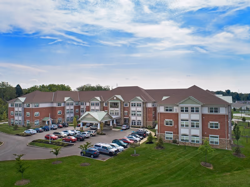 Exterior view of a large three-story senior living facility named Summit Corners with a parking lot filled with cars in front, surrounded by green lawns and trees under a partly cloudy sky.