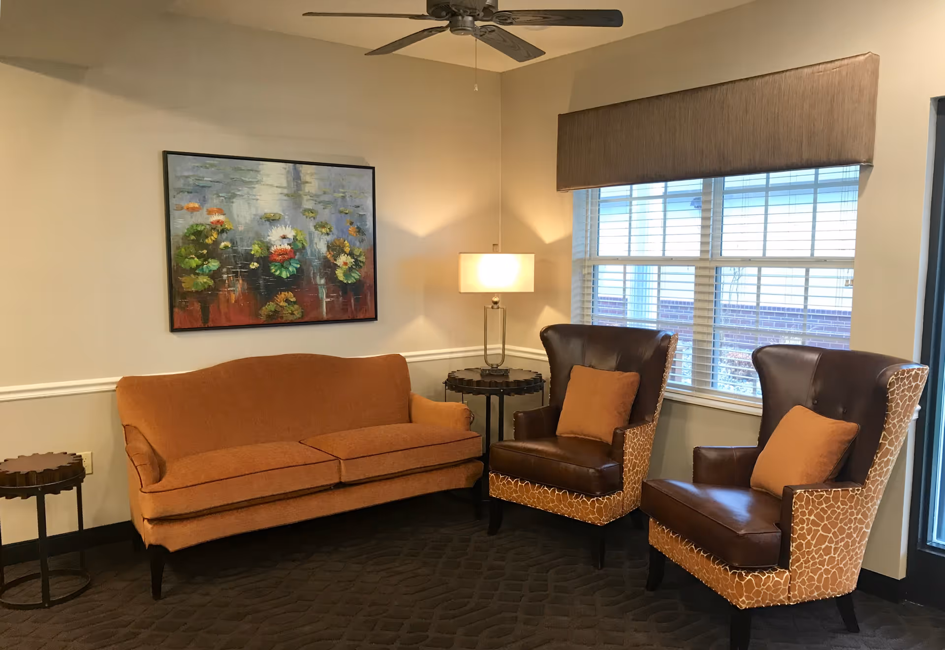 A cozy living room area with an orange sofa, two brown leather armchairs with orange cushions, a side table with a lamp, a round wooden table, and a large window with blinds. A colorful painting of water lilies hangs on the wall above the sofa.