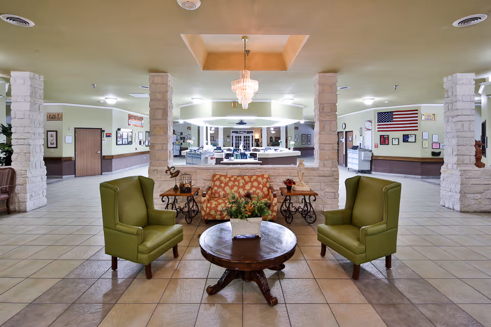 A spacious and well-lit common area in a senior living facility featuring two green armchairs and a patterned loveseat arranged around a round wooden coffee table with a potted plant. The room has tiled floors, stone pillars, and a chandelier hanging from a recessed ceiling. The background shows a reception desk and walls decorated with framed pictures and an American flag.