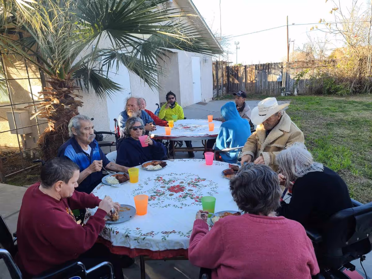 A group of elderly people sitting around two outdoor tables covered with floral tablecloths, eating a meal and drinking from colorful plastic cups. The setting is a patio area with a palm tree, adjacent to a building and a grassy yard in the background.