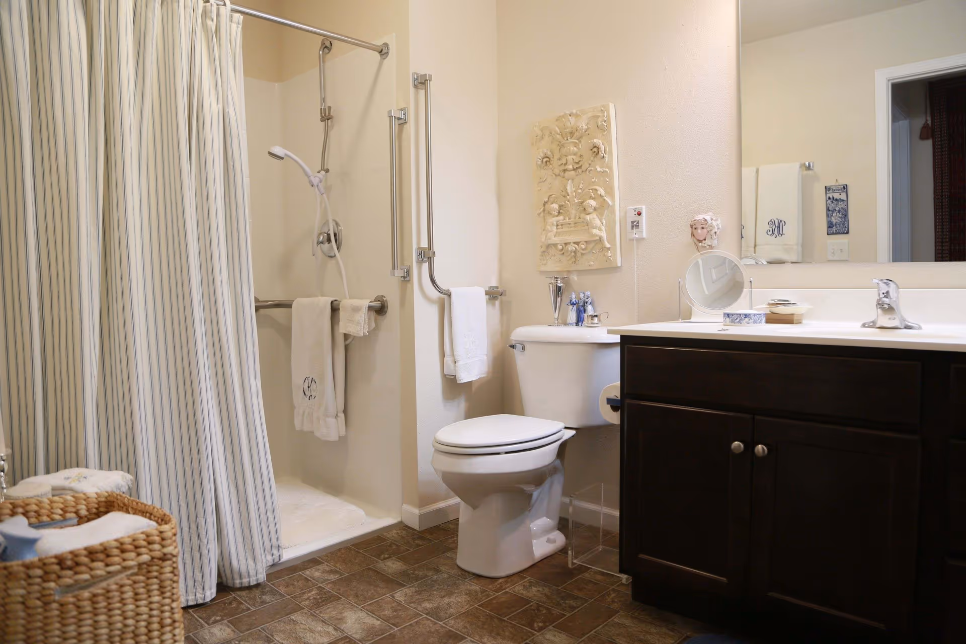 A clean bathroom featuring a shower with a striped curtain, a white toilet, and a dark wood vanity with a sink and mirror. The shower has grab bars and a handheld showerhead. Towels hang on bars near the shower and toilet. Decorative items are placed on the toilet tank and countertop. The floor has brown tiles and a woven basket with towels is visible in the corner.