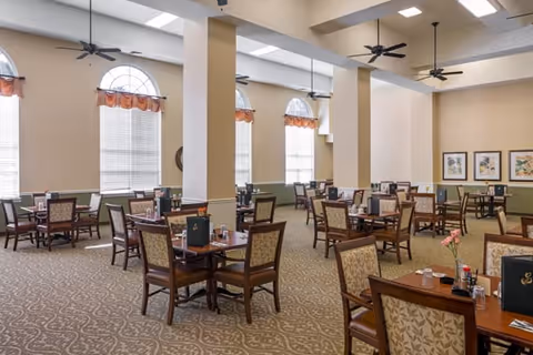 A spacious dining room with multiple wooden tables and cushioned chairs arranged neatly. The room features large arched windows with blinds and valances, ceiling fans, and soft lighting. The walls are decorated with framed artwork, and the carpet has a patterned design.