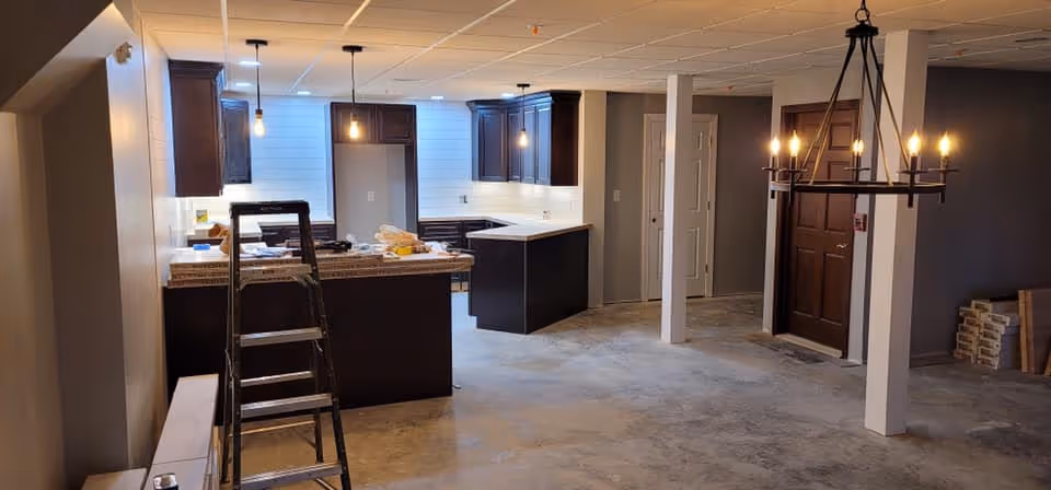 Interior view of a kitchen area under renovation with dark wood cabinets, a countertop with a ladder in front, pendant lights hanging from the ceiling, and a chandelier near a dark wooden door. The floor is unfinished concrete and there are construction materials around.