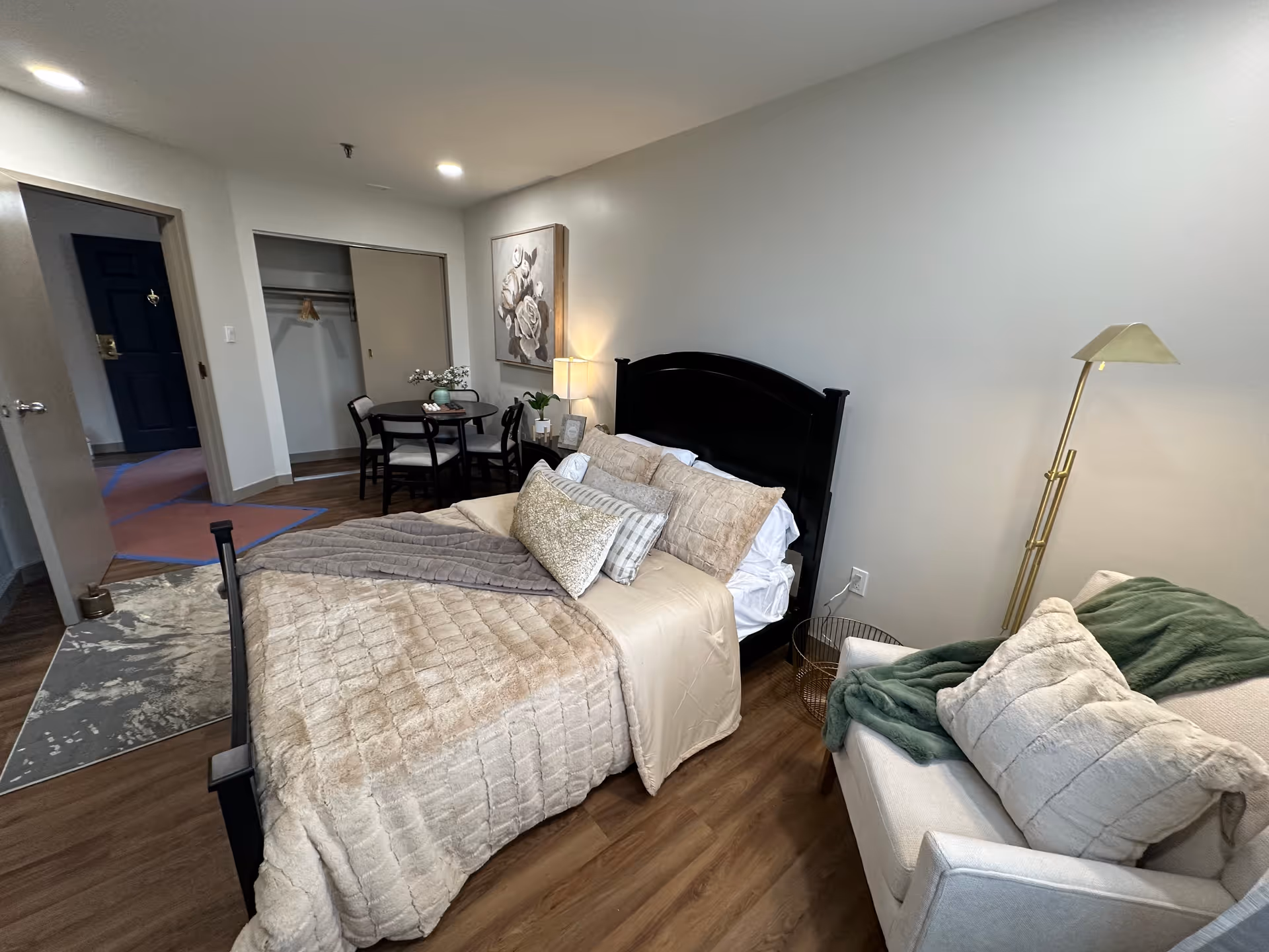 A furnished bedroom in a senior living apartment with a made bed, armchair, floor lamp and small table near an open closet.