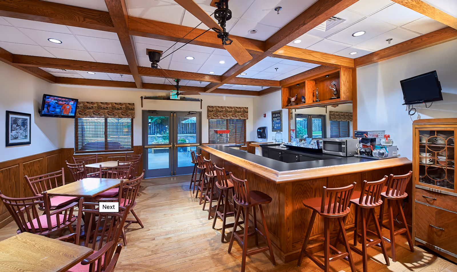 Cozy dining and bar area with a wooden counter and stools, several dining tables and chairs, and windows looking outside.