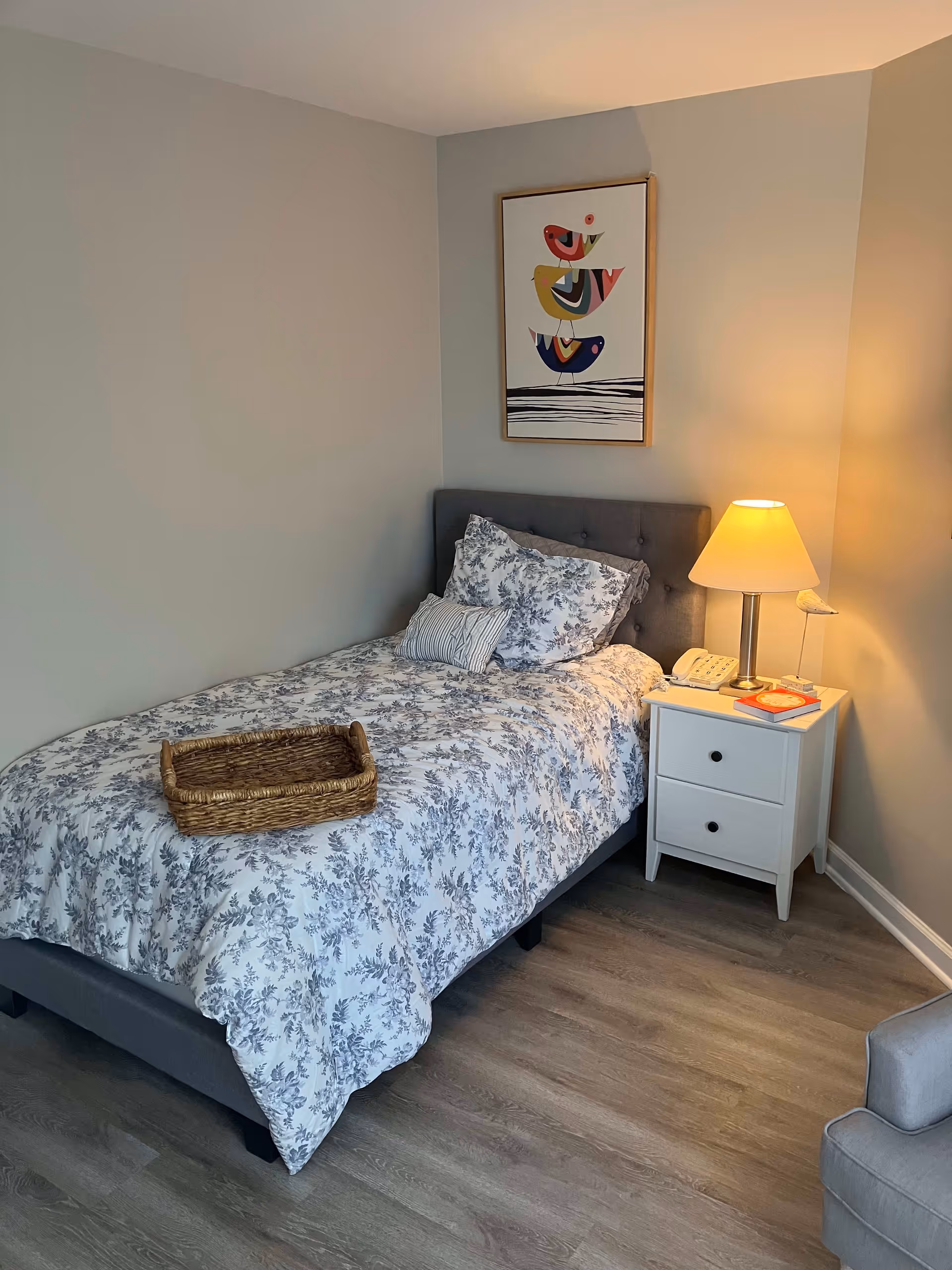 A small bedroom with a single bed covered in a white and gray floral patterned comforter and pillows. A wicker basket is placed on the bed. Next to the bed is a white nightstand with two drawers, a lamp, a telephone, and a book. Above the bed hangs a framed artwork featuring three colorful birds stacked vertically. The room has light gray walls and wood flooring.
