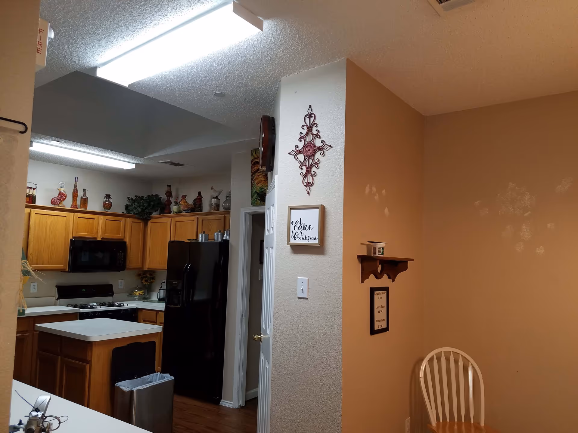 Interior view of a kitchen area with wooden cabinets, a black refrigerator, a microwave, and a stove. There is a small island with a trash bin underneath. The walls are beige with decorative items including a wall clock, a metal wall art piece, and framed signs. A wooden chair is placed against the wall with visible handprints on the wall above it.