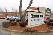 Exterior view of the Elizabeth L. Wright Apartments sign with a parking lot and a multi-story apartment building in the background. Trees and landscaping surround the sign.