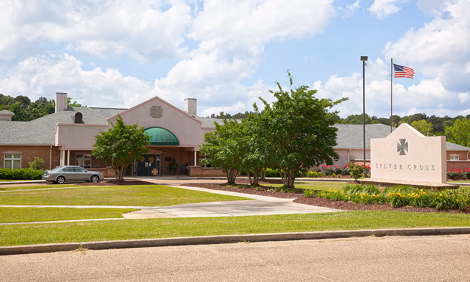 Exterior view of Silver Cross Health & Rehab facility showing a single-story building with a green awning over the entrance, surrounded by well-maintained landscaping including trees and flower beds. An American flag flies on a flagpole near a stone sign with the facility's name.