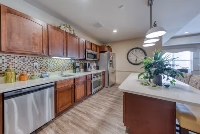 A modern kitchen with wooden cabinets, stainless steel appliances including a dishwasher, oven, microwave, and refrigerator. The kitchen features a mosaic tile backsplash, a white countertop island with a potted plant, and three pendant lights hanging above the island. The floor is wood-style, and a large clock is mounted on the wall in the background.
