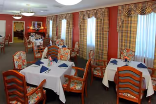 A dining room with multiple tables covered in white tablecloths and set with blue napkins. The chairs have floral upholstery and wooden frames. The room has red walls with several windows covered by plaid curtains. There are ceiling lights and a sideboard with decorative items in the background.