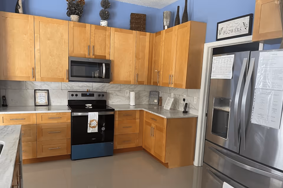 A modern kitchen with light wood cabinets, a stainless steel microwave above a black electric stove, and a stainless steel refrigerator with double doors and a bottom freezer. The countertops are light-colored, and the backsplash has a marble-like design. There are decorative items on top of the cabinets and a framed sign above the refrigerator that says 'Fresh Cut FLOWERS'.