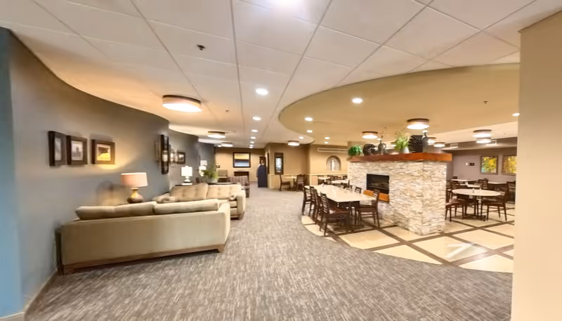 Spacious senior living common area with sofas on the left and multiple dining tables arranged around a central stone fireplace.