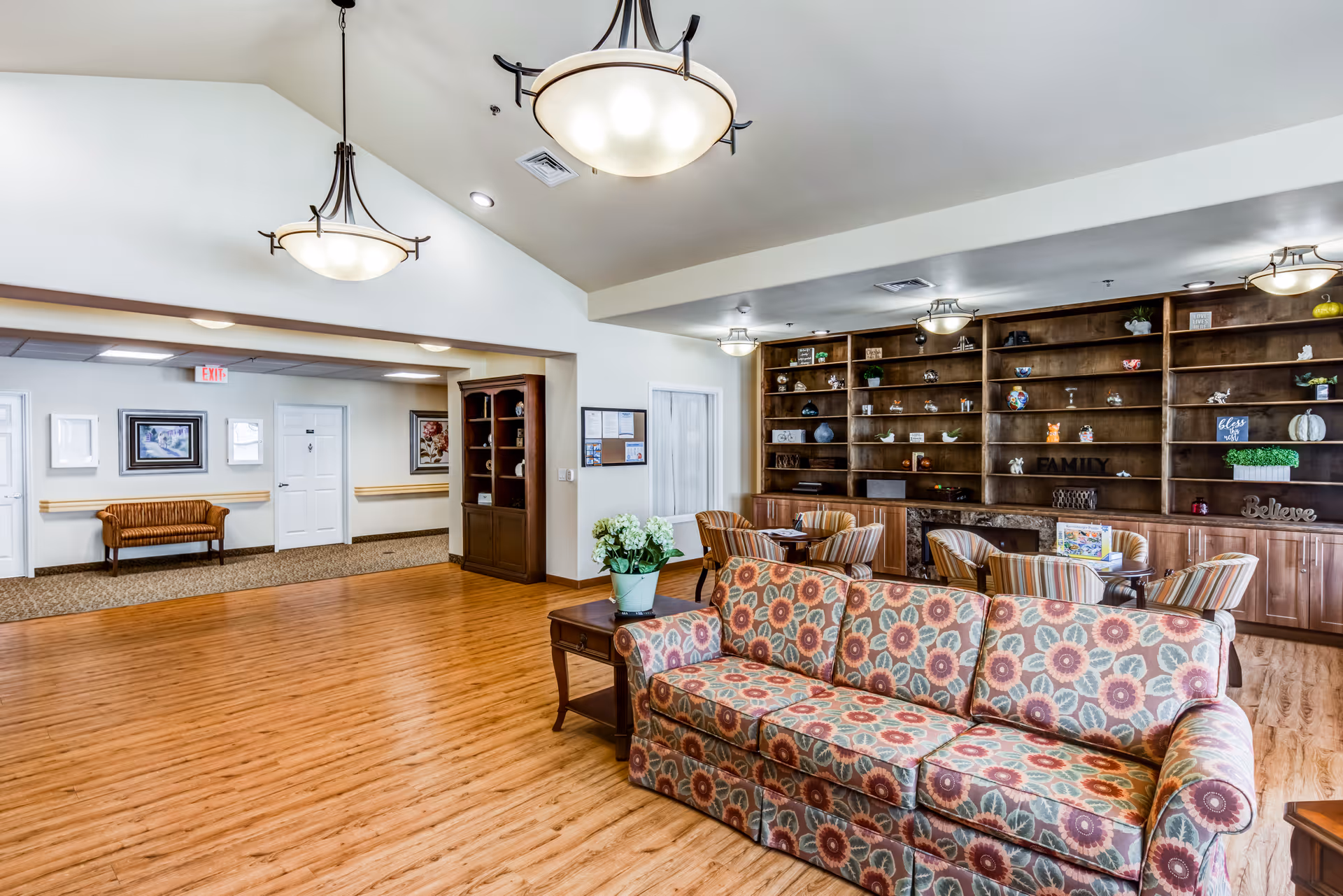 Bright senior living common room with a patterned sofa, wooden bookshelves, round tables and chairs, and hanging pendant lights.