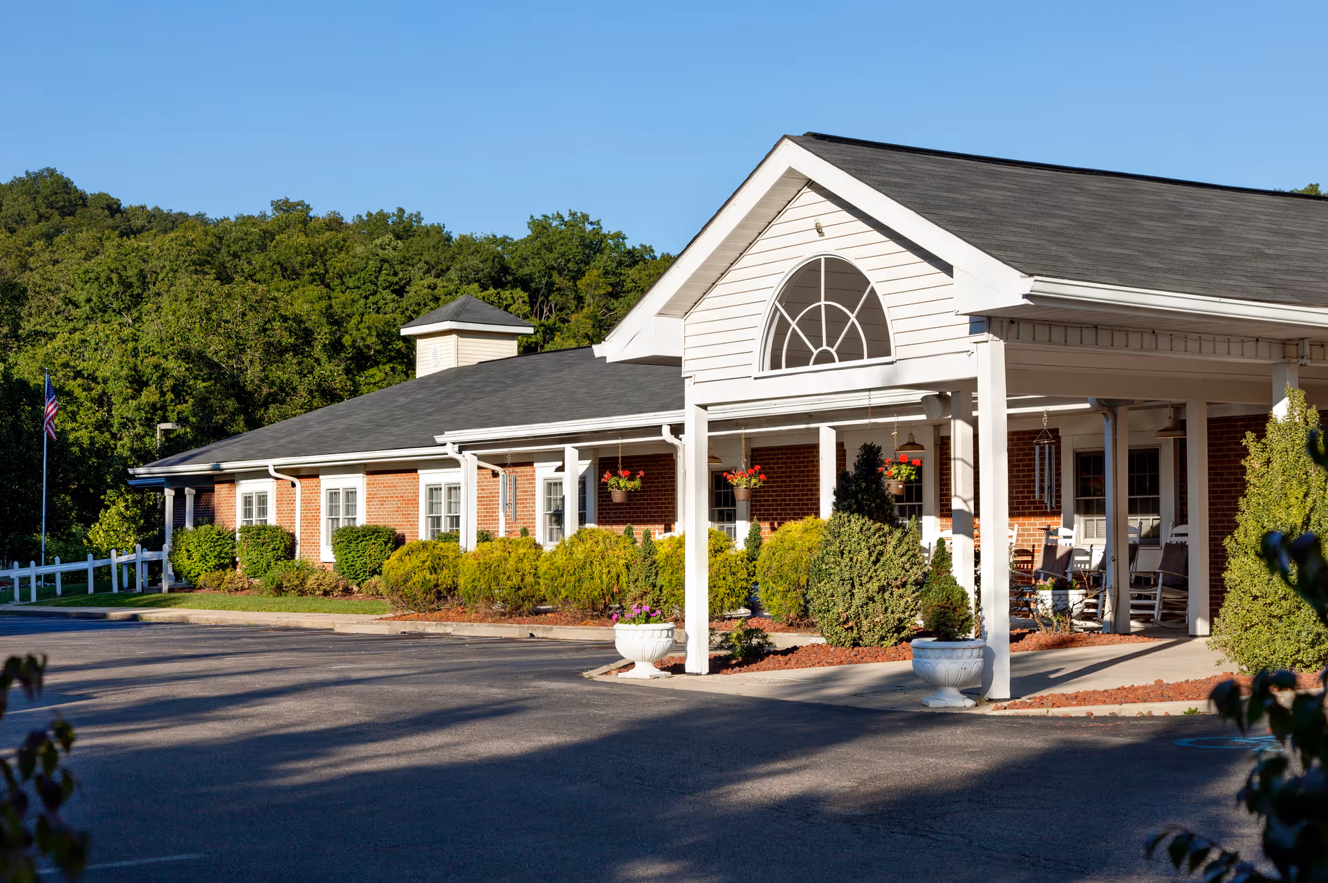 Front entrance of a brick assisted living building with a covered portico, rocking chairs, planters, and landscaped shrubs.