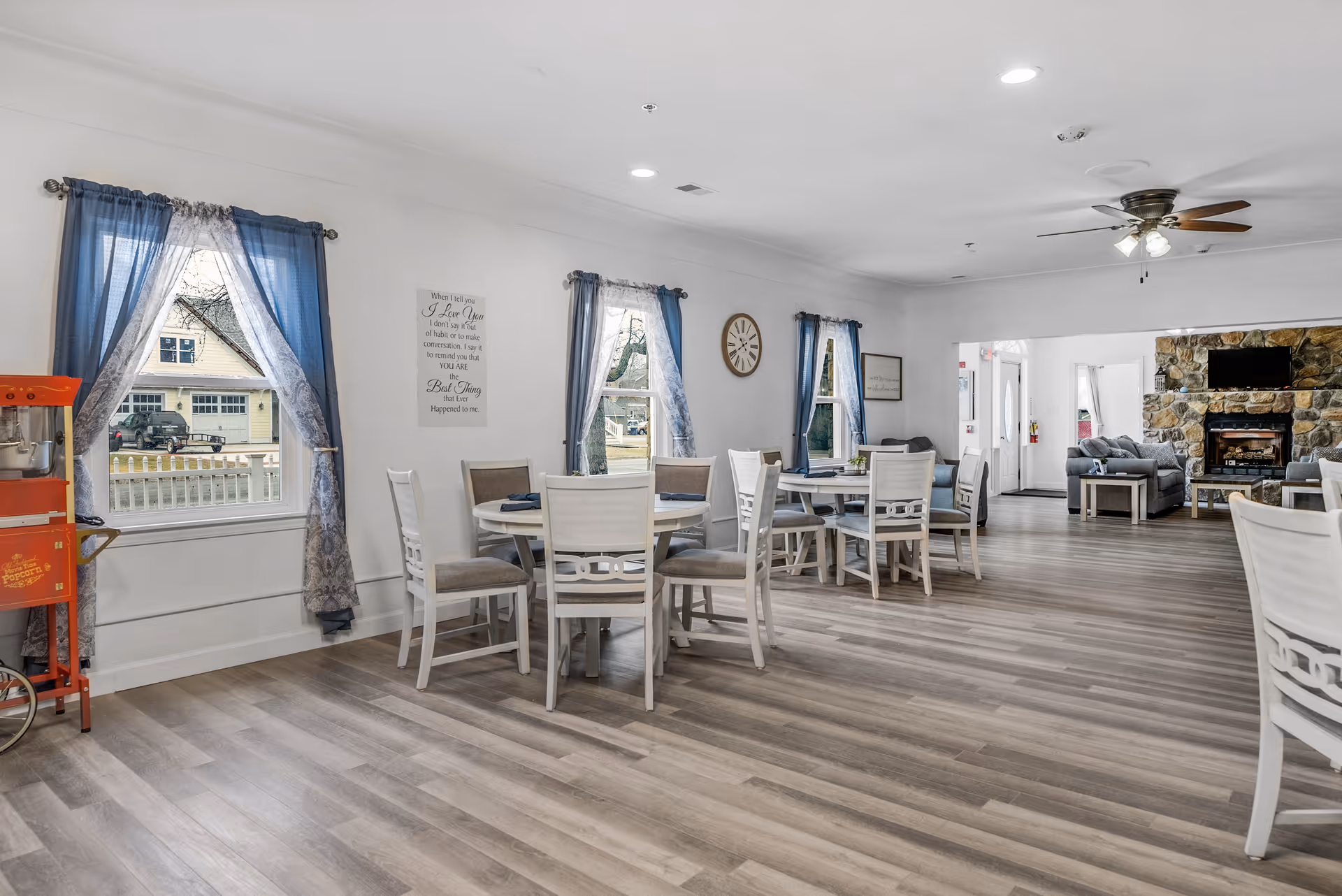 A bright and spacious common area in an assisted living facility featuring several round tables with chairs arranged around them. The room has large windows with blue curtains, light wood flooring, and white walls adorned with decorative signs and a clock. In the background, there is a cozy seating area with sofas and a stone fireplace with a mounted TV above it. A red popcorn machine is visible near one of the windows.