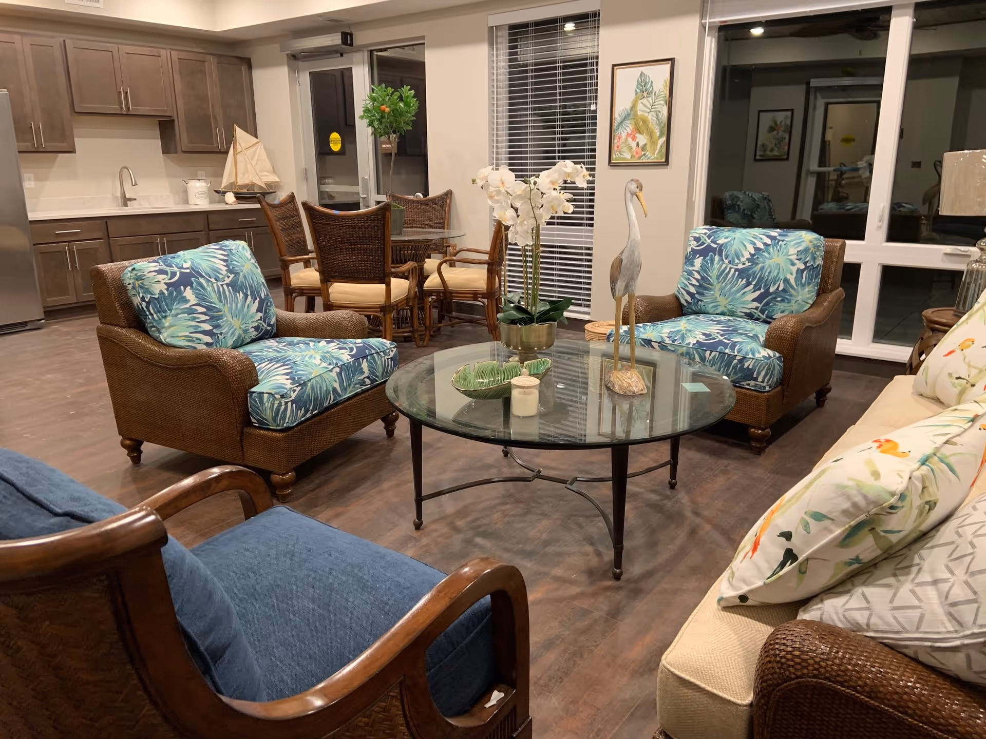 A cozy senior living common area with a glass coffee table decorated with a white orchid and a bird figurine. Surrounding the table are two armchairs with blue and green floral cushions, a blue cushioned wooden chair, and a beige sofa with floral pillows. In the background, there is a dining table with wicker chairs and a kitchen area with wooden cabinets and a stainless steel refrigerator.