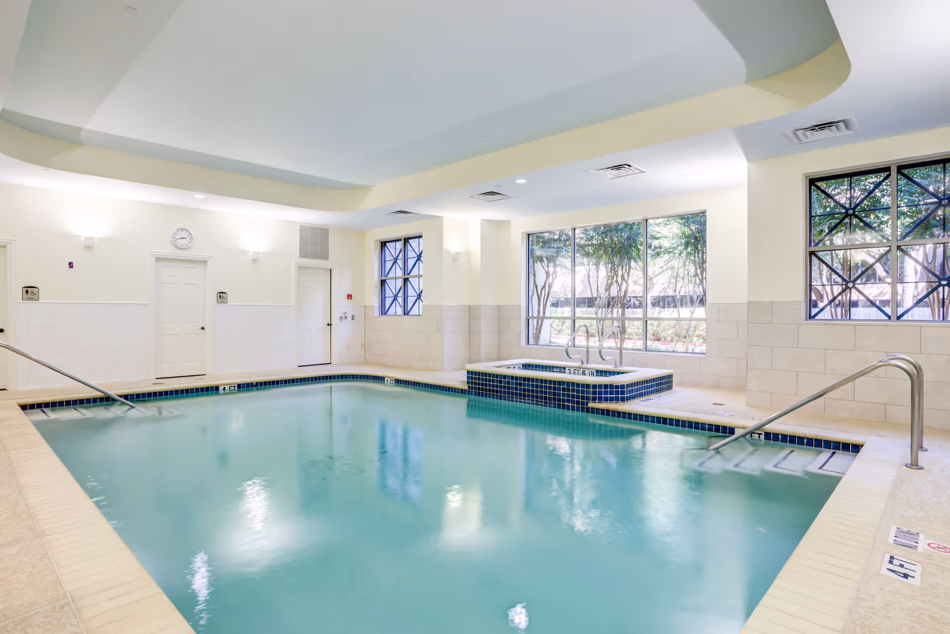 Indoor swimming pool with clear water, surrounded by beige tiled flooring and walls. There are two metal handrails for pool entry, a hot tub in the corner with blue tile accents, large windows letting in natural light, and a white ceiling with recessed lighting.