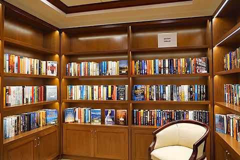 A cozy library corner with wooden bookshelves filled with a variety of books, including a section labeled 'Mystery Novels.' There is a cushioned armchair with wooden armrests positioned in front of the shelves. The shelves are well-lit with built-in lighting.