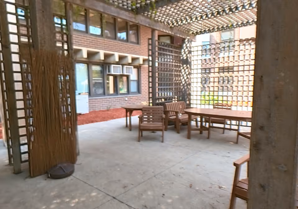 Covered outdoor patio with wooden tables and chairs under a lattice pergola next to a brick building.