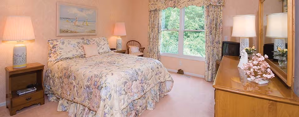A cozy bedroom with a floral-patterned bedspread on a double bed, two bedside tables each with a lamp, a wooden dresser with a mirror, a small TV, and a window with floral curtains showing greenery outside.