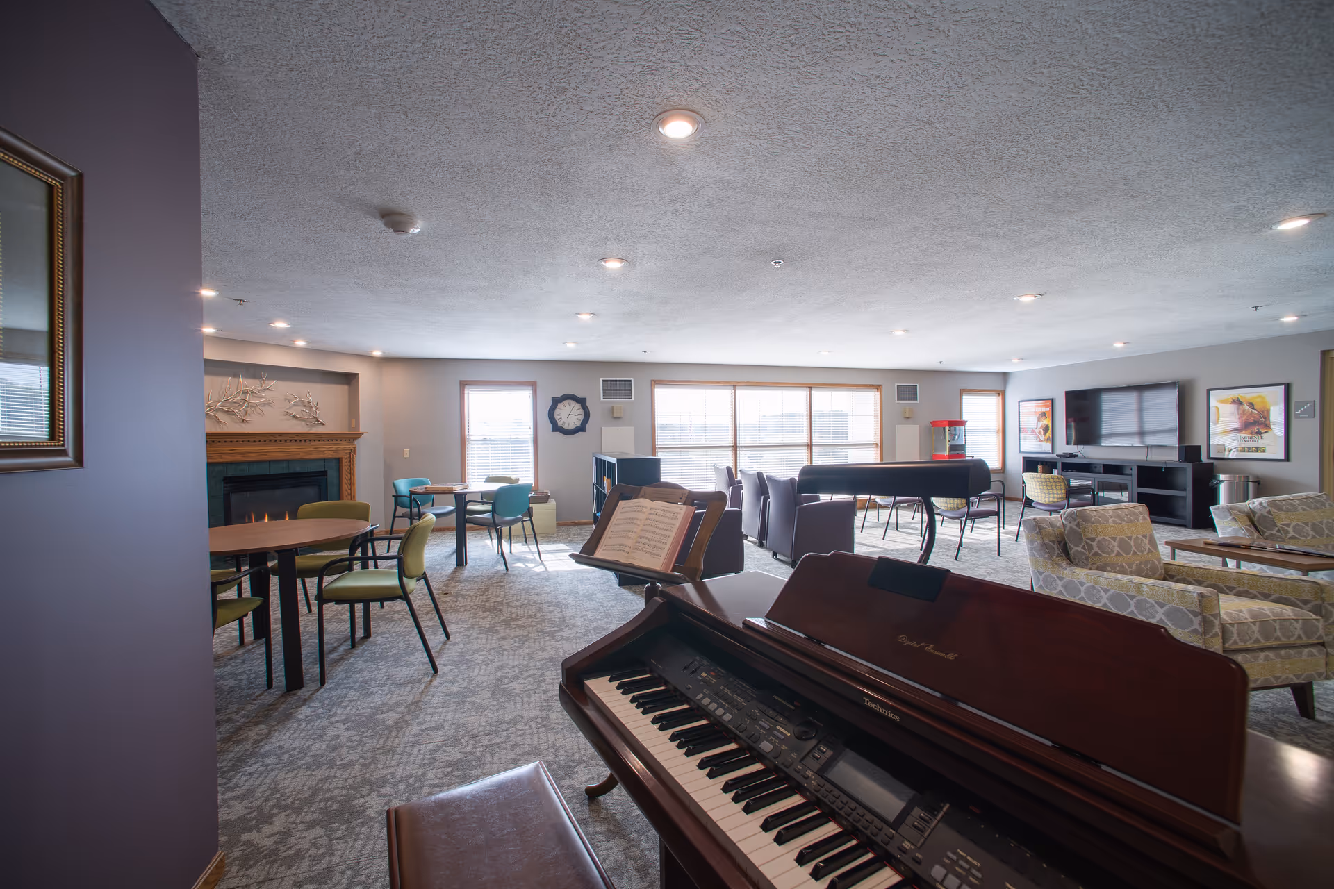 A spacious common living area with a piano in the foreground, several tables and chairs, a fireplace, large windows letting in natural light, a television mounted on the wall, and comfortable armchairs arranged for socializing.