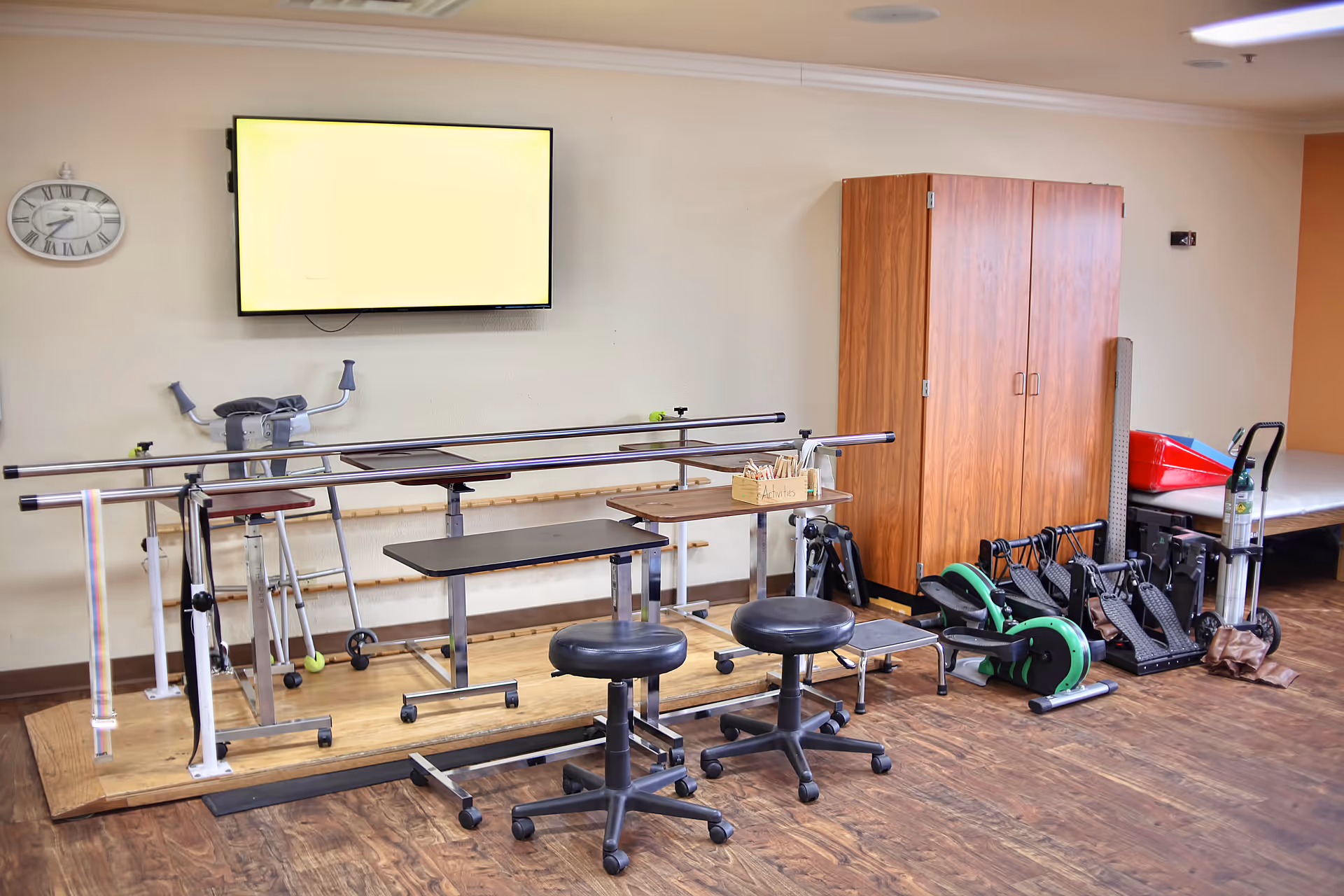 A therapy or rehabilitation room in a senior living facility with parallel bars for walking exercises, two black rolling stools, exercise equipment including pedal machines, a wooden cabinet, a wall-mounted TV, and a clock on the wall.