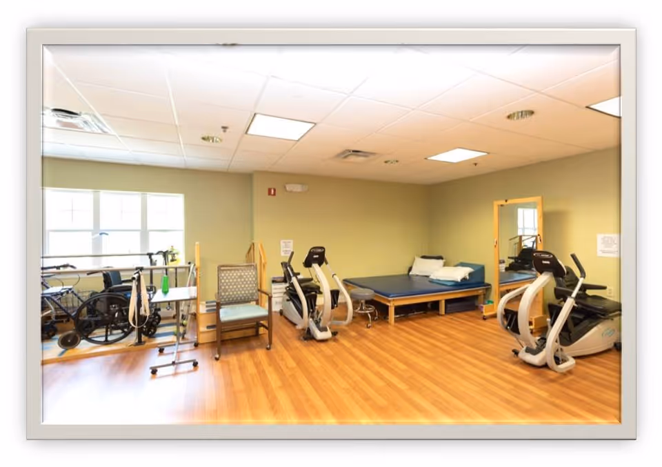 A rehabilitation or physical therapy room with exercise equipment including stationary bikes, a wheelchair, a walker, a padded therapy table with pillows, a chair, a mirror, and wooden flooring under bright ceiling lights.