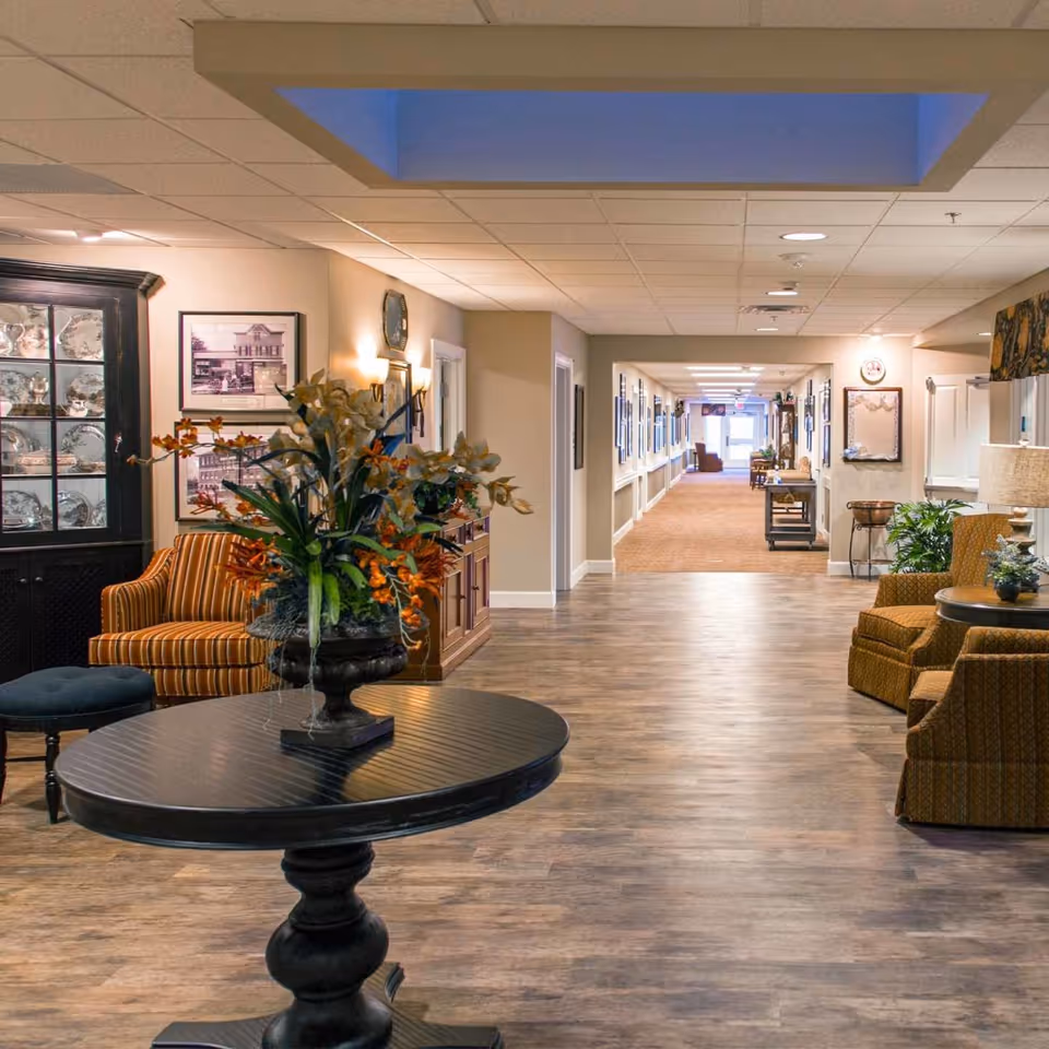 A well-lit hallway in a senior living facility with wood flooring and beige walls. The hallway features comfortable seating areas with patterned armchairs and a round black table with a floral arrangement in the foreground. The walls are decorated with framed pictures and there is a glass-front cabinet displaying dishes on the left side. The hallway extends into the distance with more seating and decor visible.