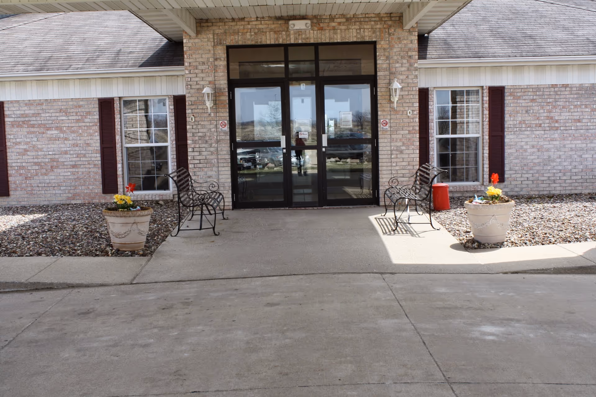Front entrance of a brick building with double glass doors, two metal benches, and potted flowers on either side.