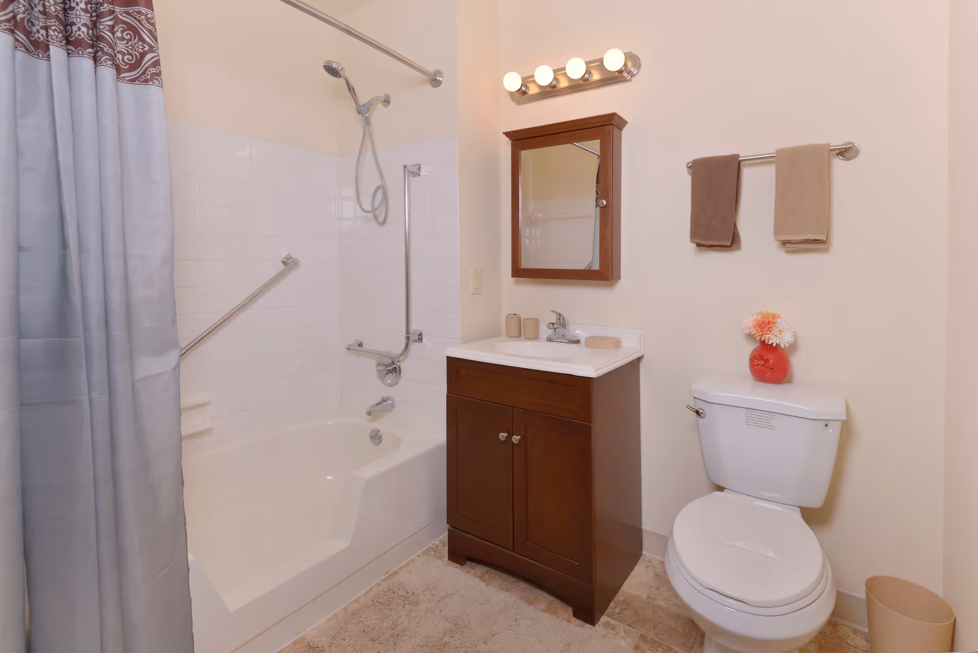 A clean bathroom with a bathtub and shower combination featuring grab bars, a shower curtain, a wooden vanity with a sink and mirror above it, a toilet with a red vase holding flowers on the tank, two towels hanging on a towel rack, and a beige trash bin on the floor.