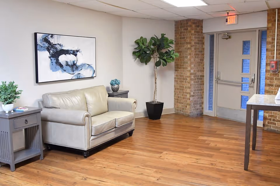 A small seating area with a beige leather loveseat, two gray side tables with plants and a small decorative item, a large abstract painting on the wall, a tall potted plant, and a door with an exit sign above it. The floor is wooden and the walls are a mix of white and exposed brick.