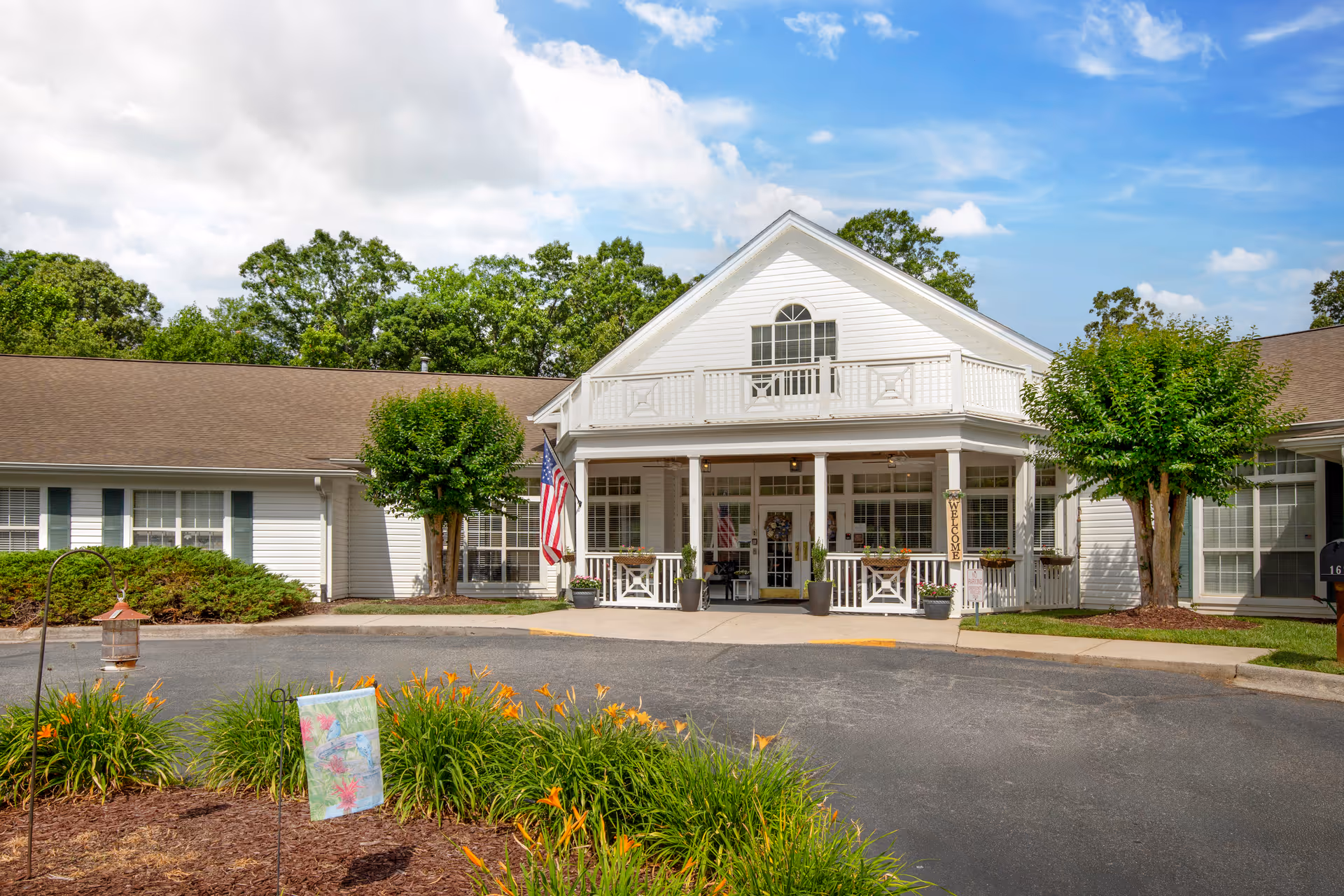 White two-story senior living building with a covered porch entrance, American flag, landscaping and a circular driveway.