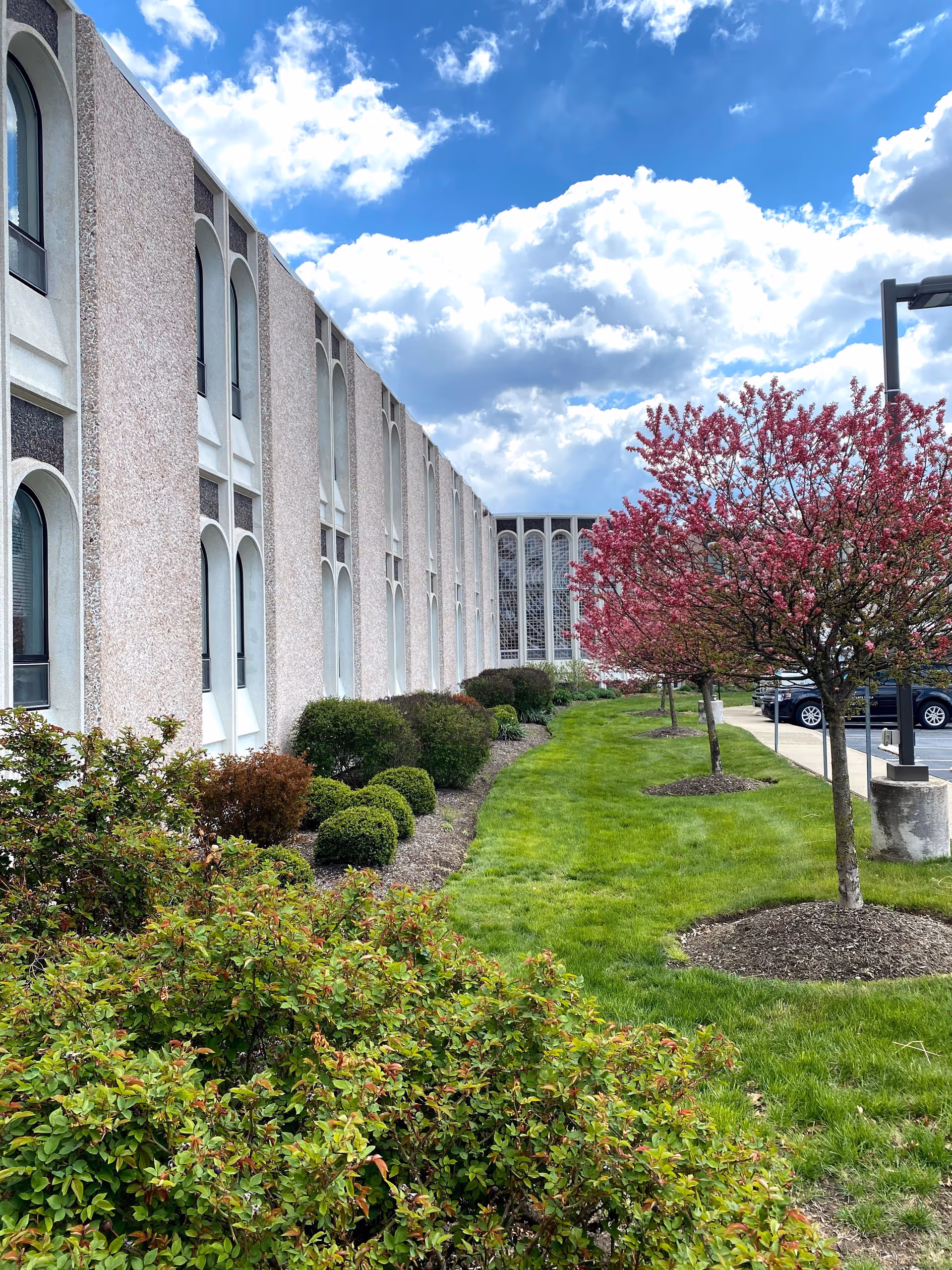 Exterior view of a senior living facility building with a row of arched windows. In front of the building, there is a well-maintained garden with green shrubs and small trees with pink blossoms. A parking lot with cars is visible on the right side under a partly cloudy blue sky.